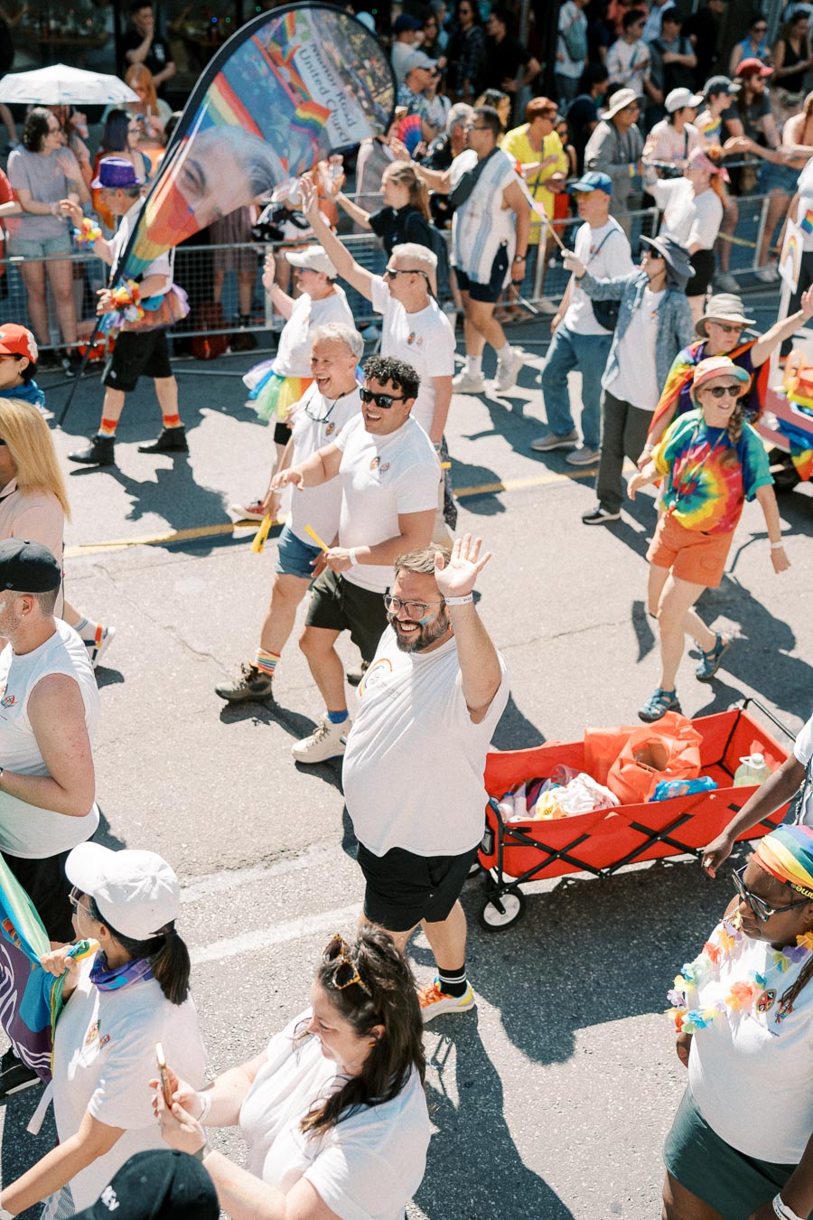 Participants in a pride parade joyfully march down the street, waving flags and wearing colorful attire. A red wagon filled with supplies is visible among the crowd.