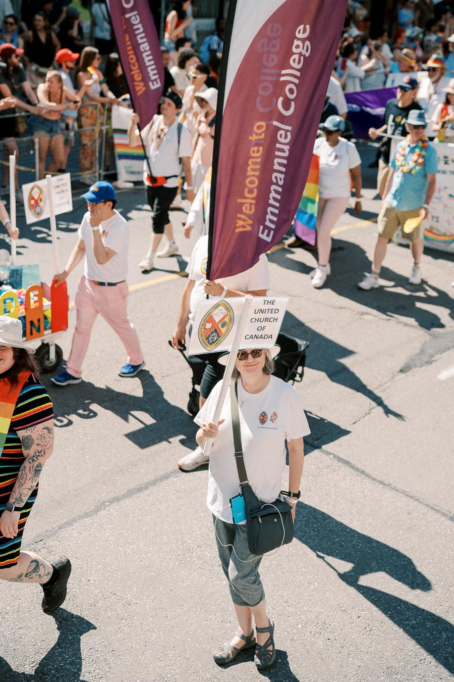 Participants in a parade hold signs for Emmanuel College and The United Church of Canada, celebrating diversity and inclusion.