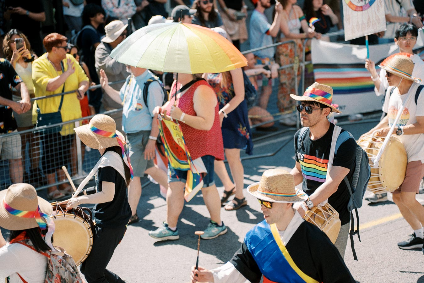 Participants in a colorful parade, wearing rainbow-themed clothing and accessories, play drums and engage with the crowd.