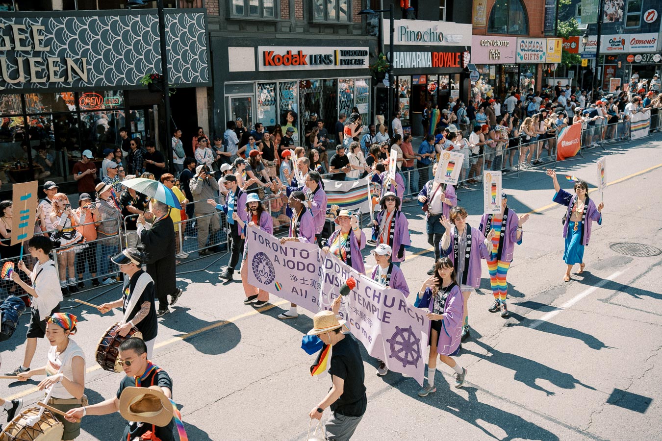 Participants from the Toronto Buddhist Church march in a parade, wearing purple robes and carrying a banner. Spectators watch from the sidewalk, with shops visible in the background.