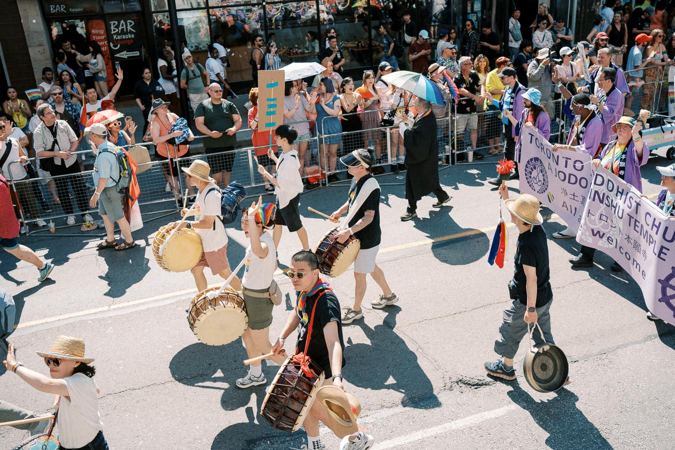 Participants in a parade play drums and carry a banner for the Toronto Buddhist Church, while spectators watch from the sidelines.