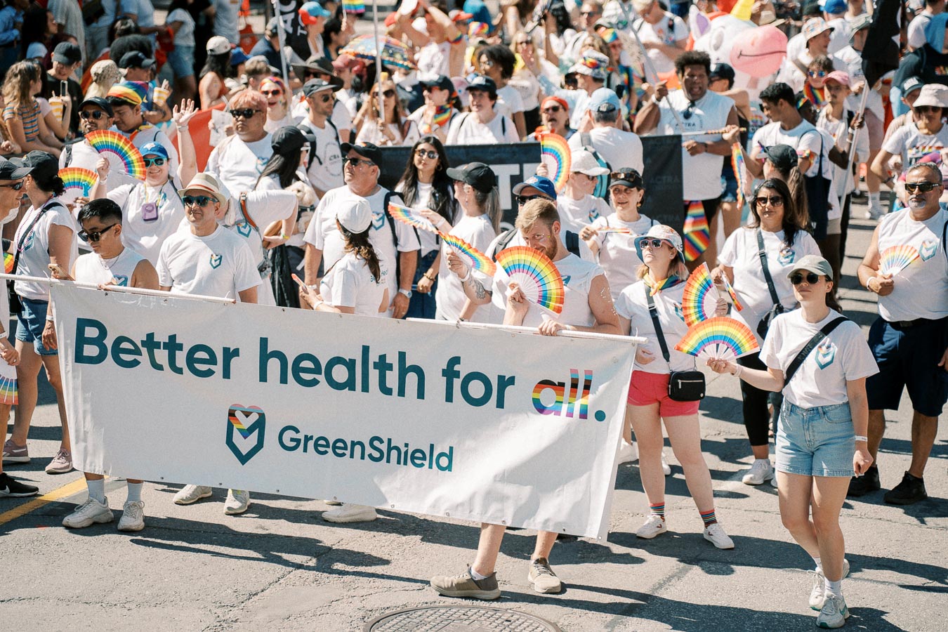 Participants in a parade hold a 'Better health for all. GreenShield.' banner, celebrating diversity and inclusion with rainbow fans.