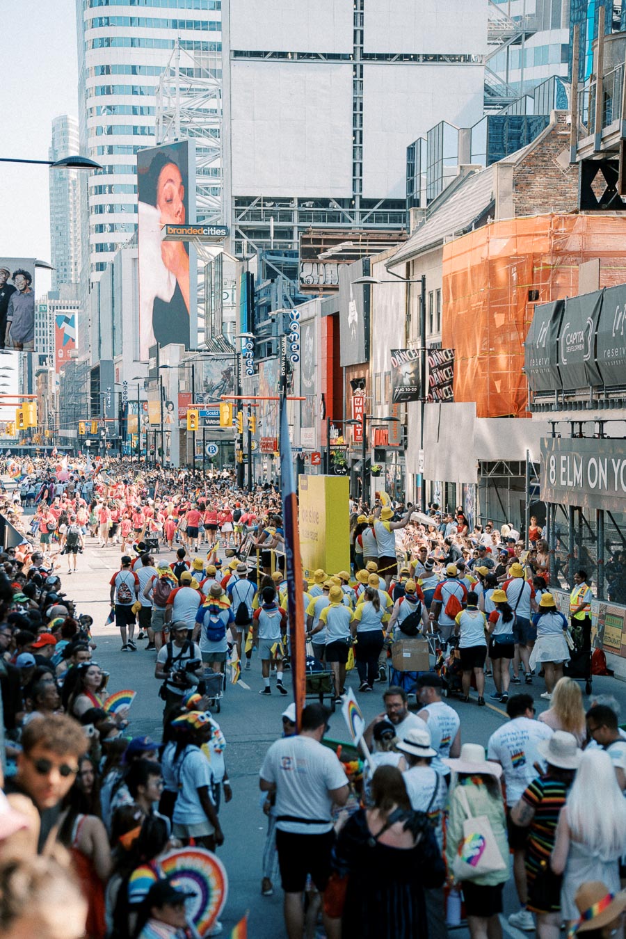 A lively parade takes place on a bustling city street, with participants in colorful attire and spectators waving rainbow flags, set against a backdrop of urban architecture and digital displays.