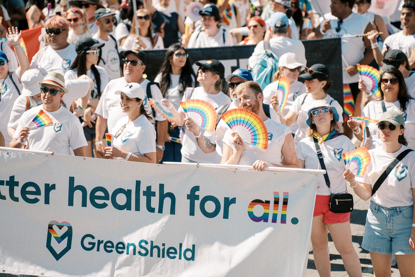 Participants at a pride parade hold rainbow fans and a banner promoting health inclusivity by GreenShield.