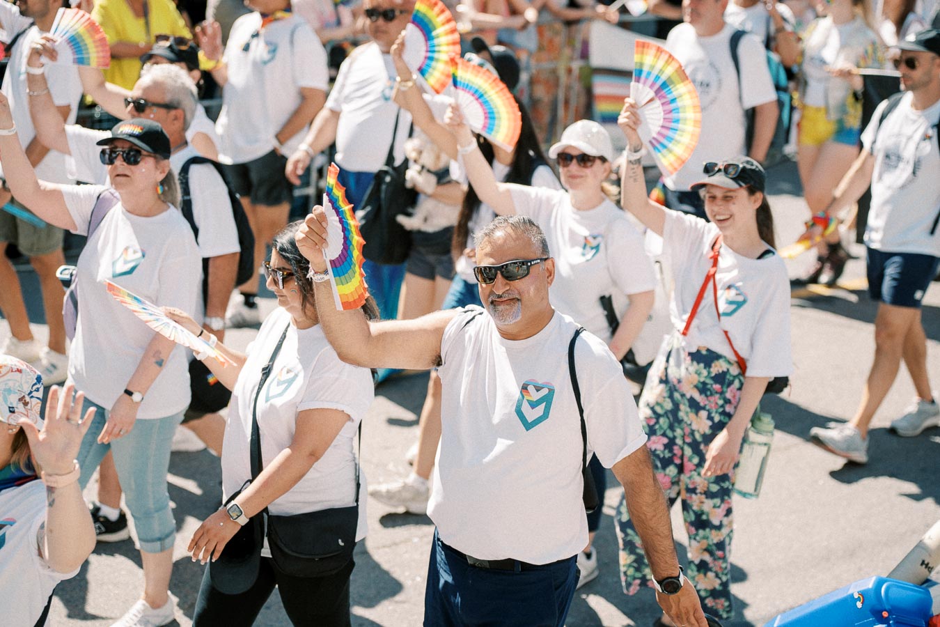 Participants in a pride parade wearing white shirts with heart designs, waving rainbow fans.