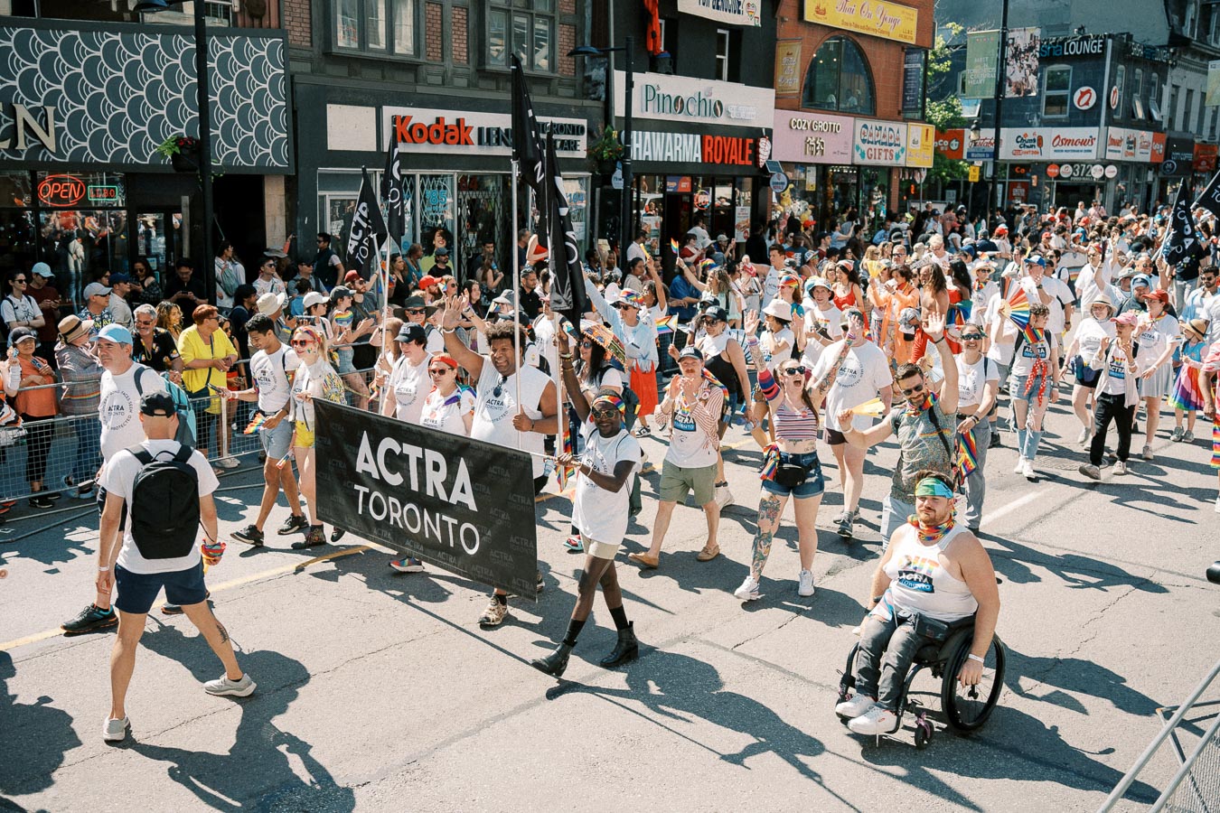 Participants in a parade march down a street in Toronto, holding an 'ACTRA Toronto' banner, surrounded by a lively crowd and colorful storefronts.