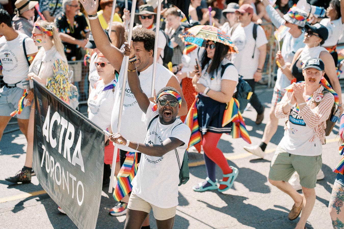 Participants in a parade march with an 'ACTRA Toronto' banner, celebrating with rainbow-themed accessories and joyful expressions.
