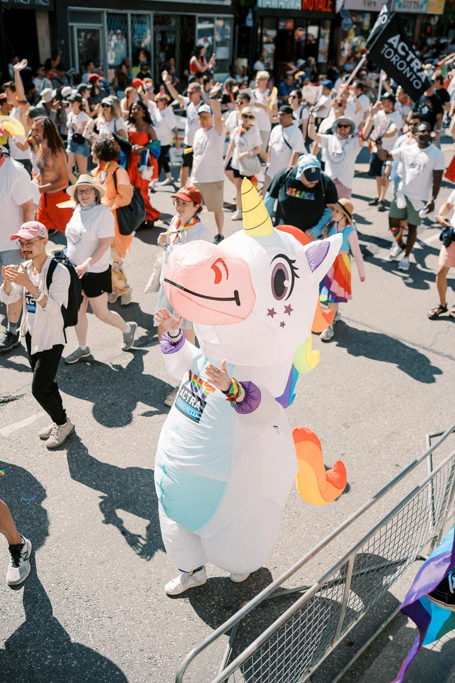 A vibrant parade scene featuring a person in a unicorn costume among a crowd celebrating with rainbow-themed accessories.