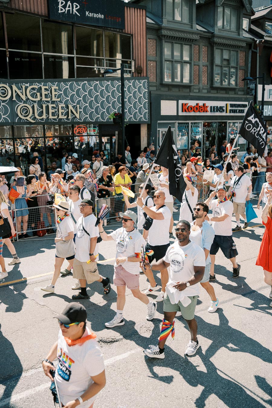 Participants march in a parade, holding flags and wearing matching T-shirts, as spectators watch from the sidelines.