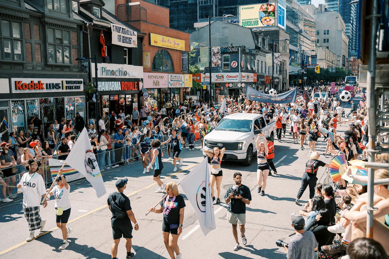 A vibrant street parade featuring participants with flags and banners, a white truck, and enthusiastic spectators lining the sidewalk.