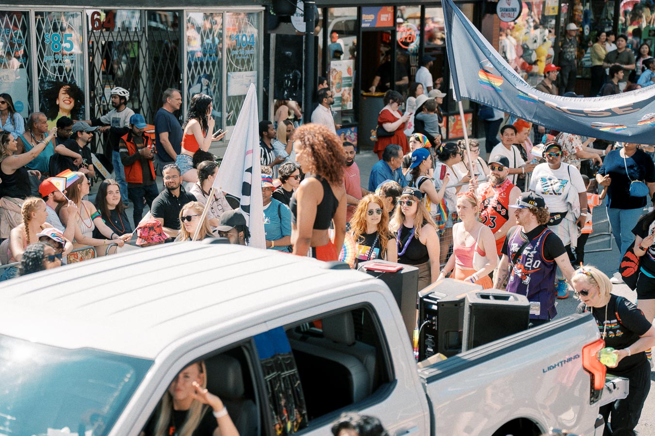 A vibrant pride parade with a diverse crowd celebrating with flags and colorful attire, featuring a white truck in the foreground.