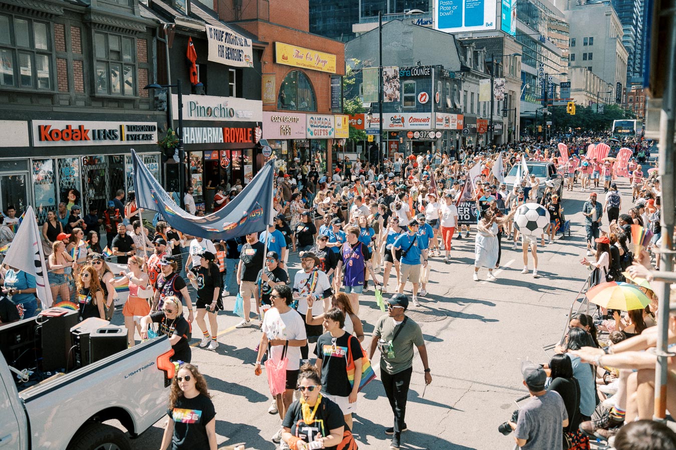 A lively parade takes place on a bustling city street, featuring participants in colorful attire and rainbow-themed accessories. The event draws a large crowd, with spectators lining the street and shops in the background.