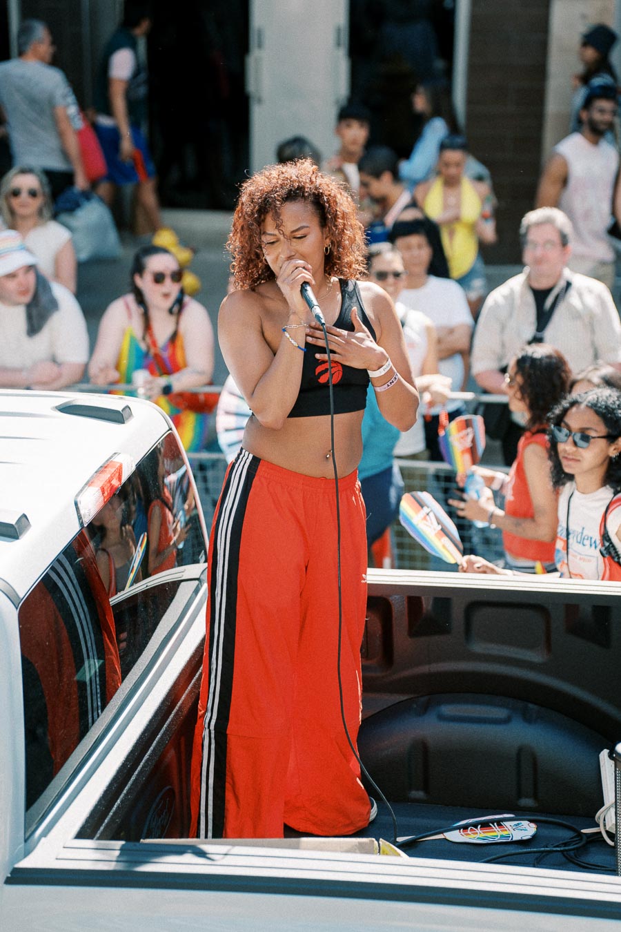 A performer sings from the back of a pickup truck during a parade, surrounded by a crowd with rainbow flags.