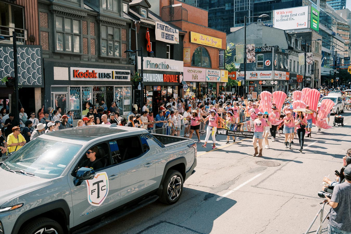 A lively parade with participants in pink outfits marches down a city street, watched by a large crowd. A gray truck leads the procession.