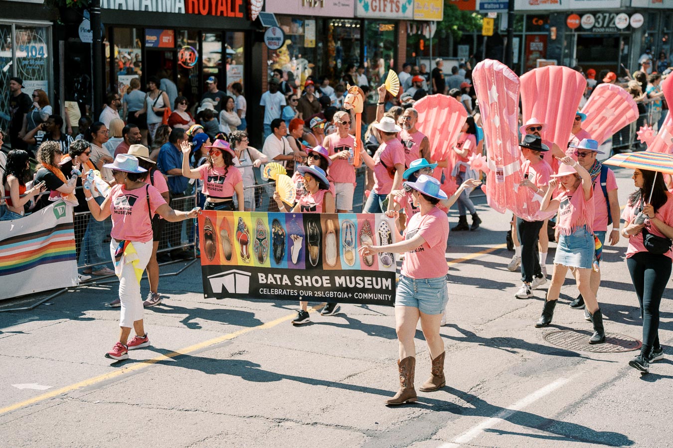 Participants from the Bata Shoe Museum march in a parade celebrating the 2SLGBTQIA+ community, wearing pink shirts and colorful hats.
