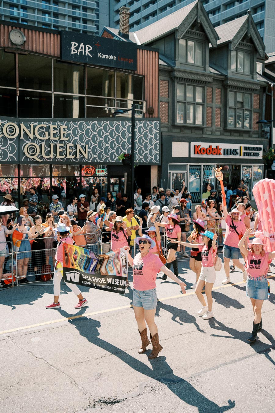 Participants in a parade celebrate with a banner for the Bata Shoe Museum, wearing pink shirts and hats, as spectators watch from the sidewalk.