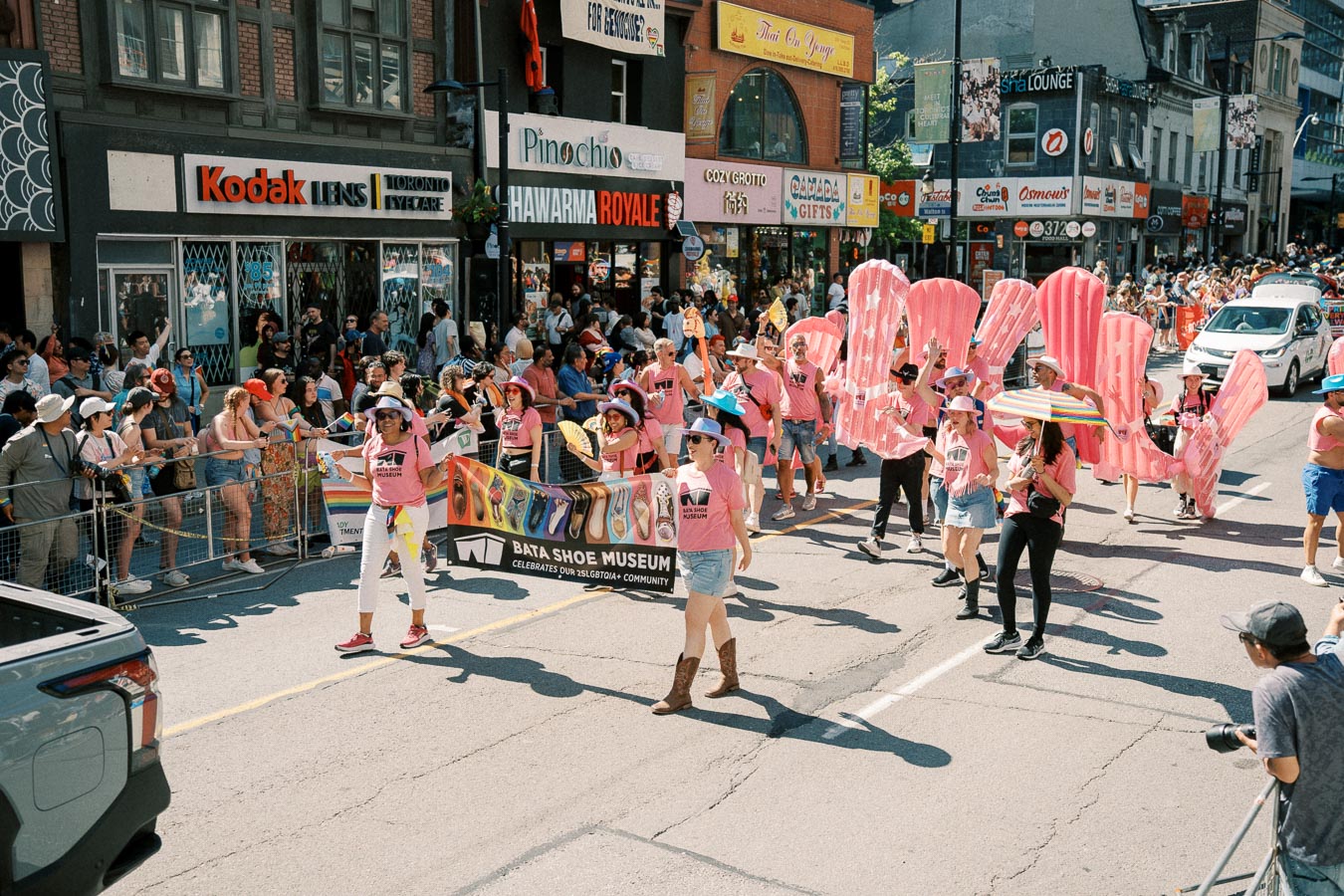 Participants in a parade, representing the Bata Shoe Museum, walk down a busy street lined with spectators and shops.