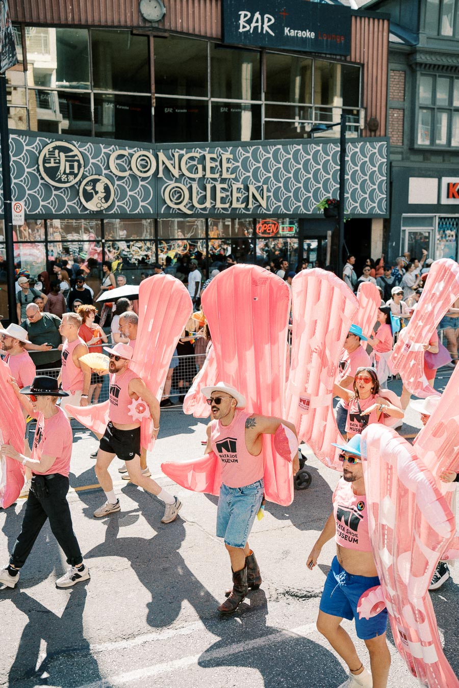Parade participants in pink attire carry large inflatable objects, passing by Congee Queen restaurant.