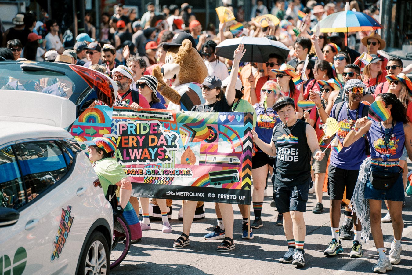Participants in a Pride parade hold a colorful banner reading 'Pride Every Day. Always.' The crowd is lively, with many wearing rainbow-themed clothing and accessories.
