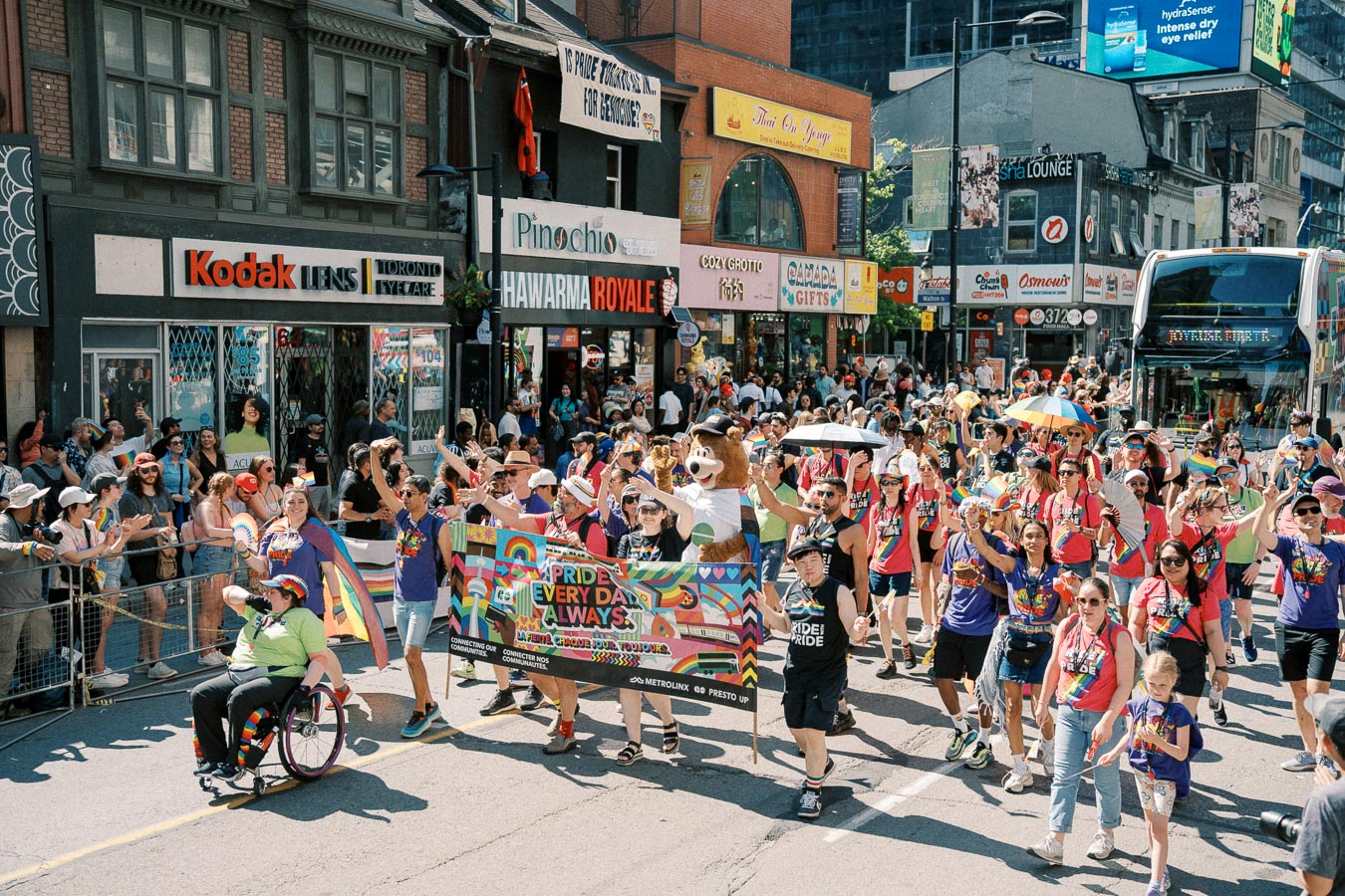 Participants in a Pride parade march down a bustling street, celebrating with colorful banners and rainbow attire. Spectators line the sidewalks, cheering and capturing the moment.
