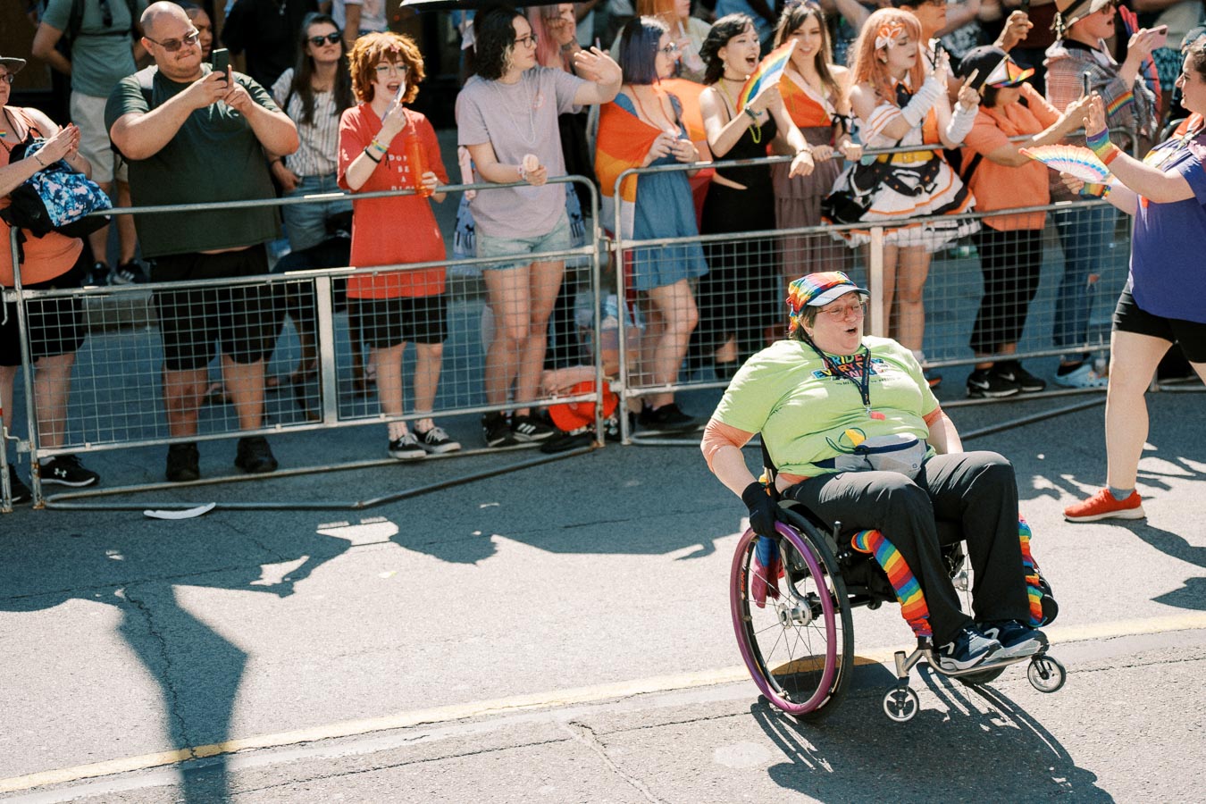 A participant in a wheelchair, adorned in rainbow colors, takes part in a pride parade, cheered on by a diverse crowd.