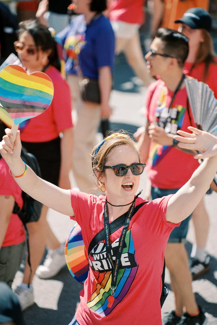 A participant in a pride parade wearing a pink shirt with a rainbow design, holding a rainbow heart sign, and surrounded by others in colorful attire.
