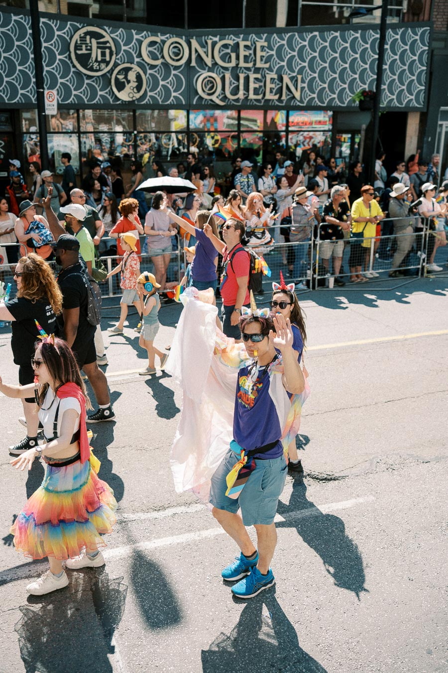 Participants in a pride parade wearing vibrant outfits and accessories, with spectators gathered behind barriers. The 'Congee Queen' restaurant sign is visible in the background.