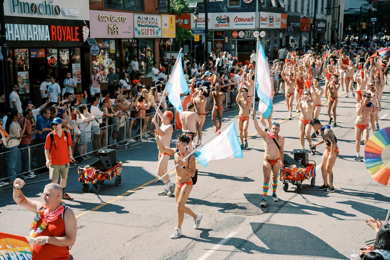 Participants in a pride parade march down a city street, waving transgender flags and wearing red swimwear, as a crowd cheers them on.