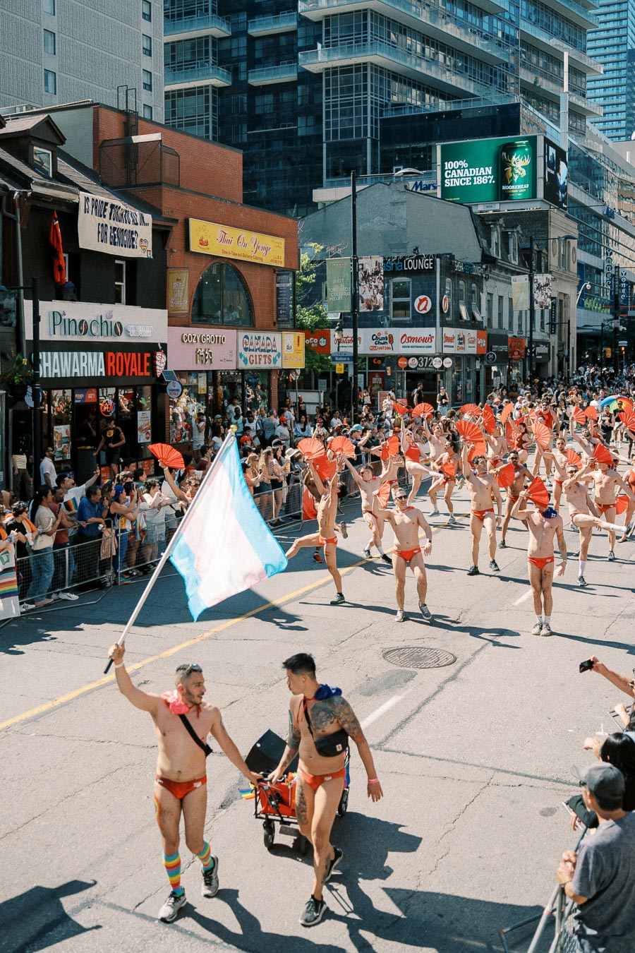 Participants in a colorful parade march down a city street, waving flags and wearing red outfits, while spectators line the sidewalks.