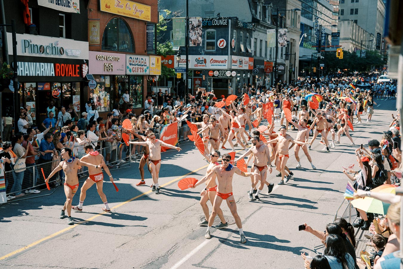 Participants in red swimwear and fans march in a vibrant parade, cheered on by a lively crowd in a bustling city street.