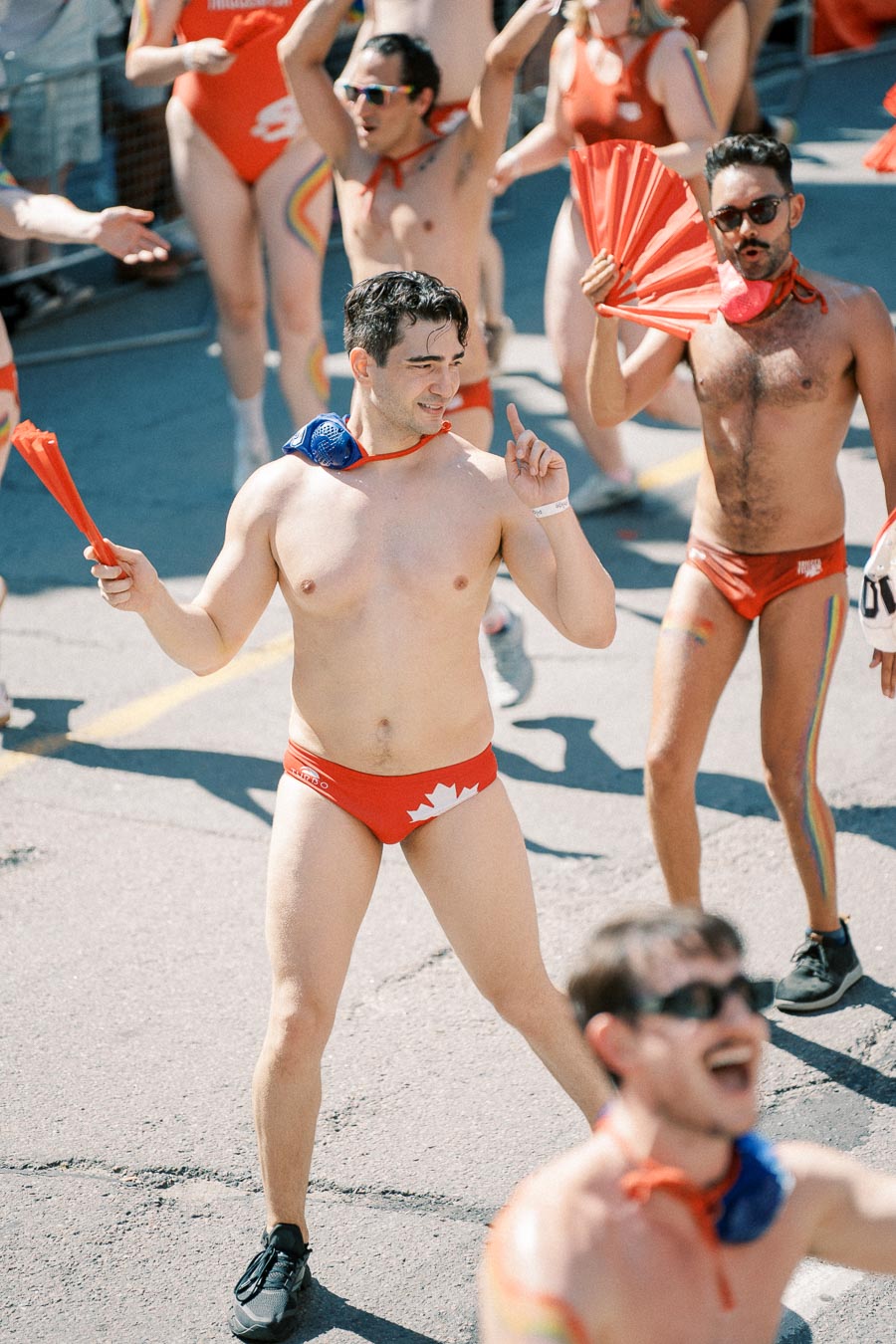 Participants in a parade wearing red swimwear with Canadian flag motifs, holding red fans, and celebrating with enthusiasm.