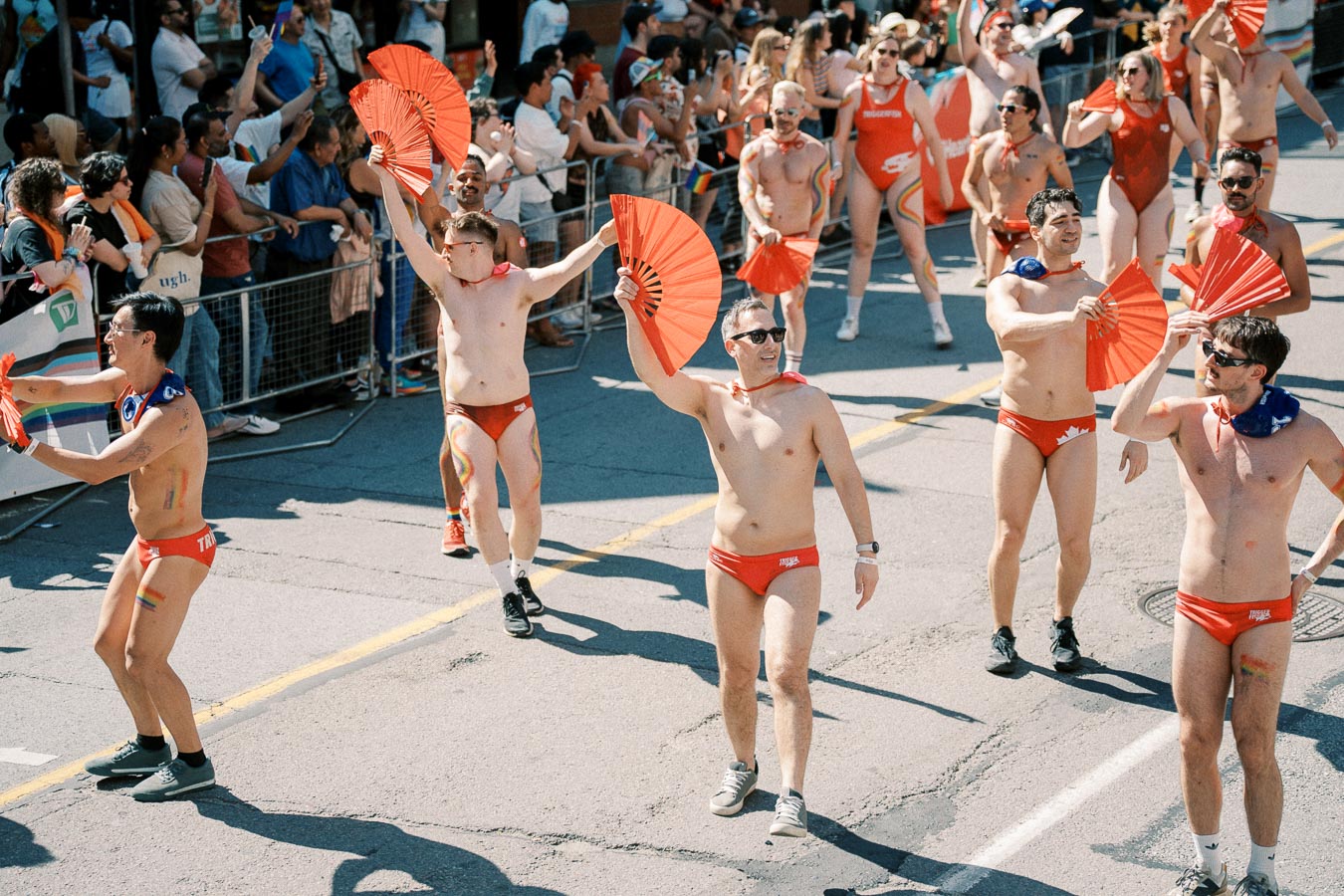 Participants in red swimwear and capes wave red fans during a lively parade, with spectators lining the street.