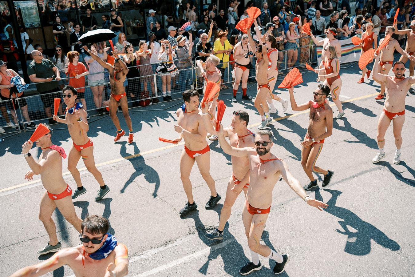 Participants in vibrant red swimwear and accessories march joyfully in a parade, waving red fans, as spectators cheer from the sidelines.