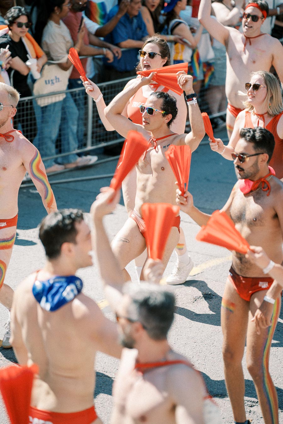 Participants in a pride parade celebrate with red outfits and fans, showcasing rainbow body paint and accessories.
