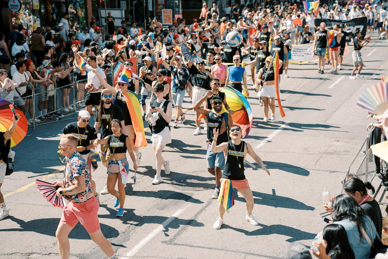 Participants in a lively pride parade march down a city street, celebrating with rainbow flags and umbrellas, while spectators cheer from the sidelines.