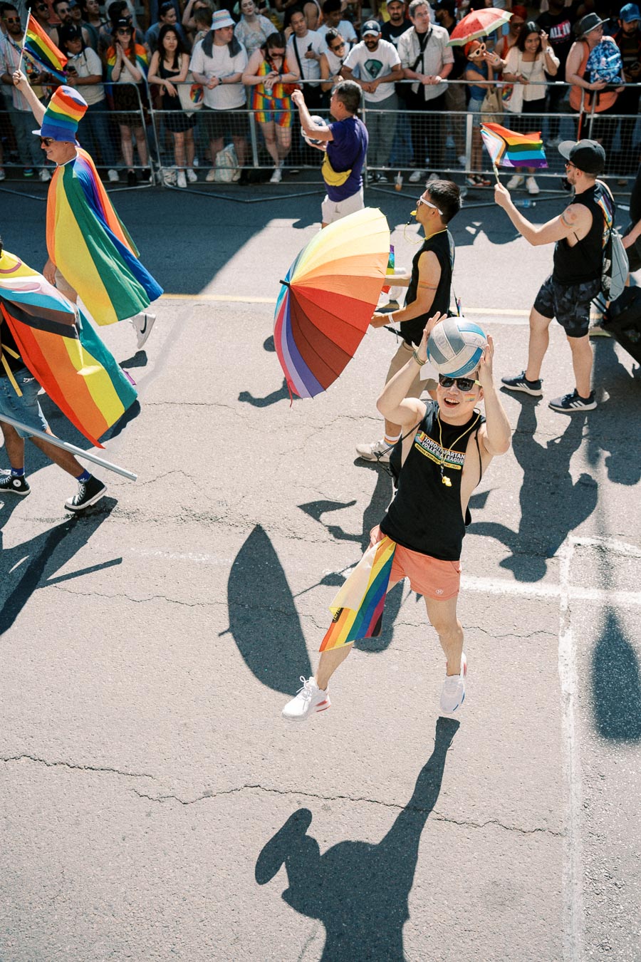 Participants in a pride parade celebrate with rainbow flags and umbrellas, while a crowd watches from behind a barrier.