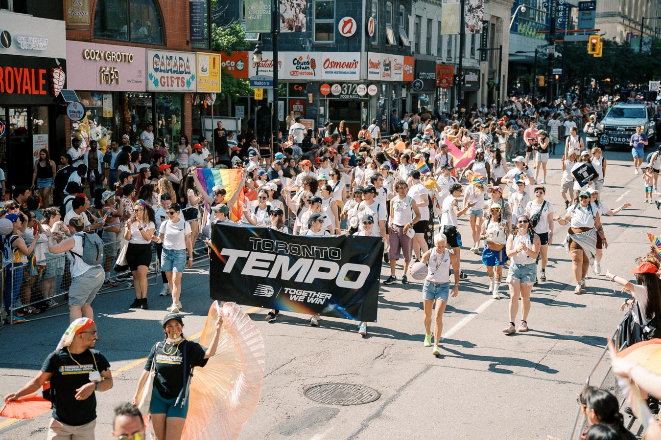 Participants in a Toronto parade march with a 'Toronto Tempo' banner, celebrating diversity and inclusion with rainbow flags and festive attire.