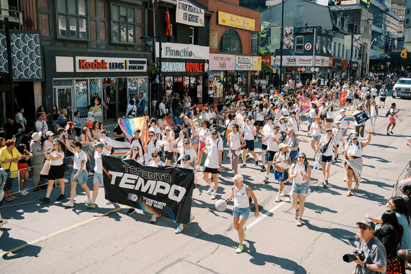 Participants in a parade march down a city street, holding a "Toronto Tempo" banner and rainbow flags, celebrating diversity and inclusion.