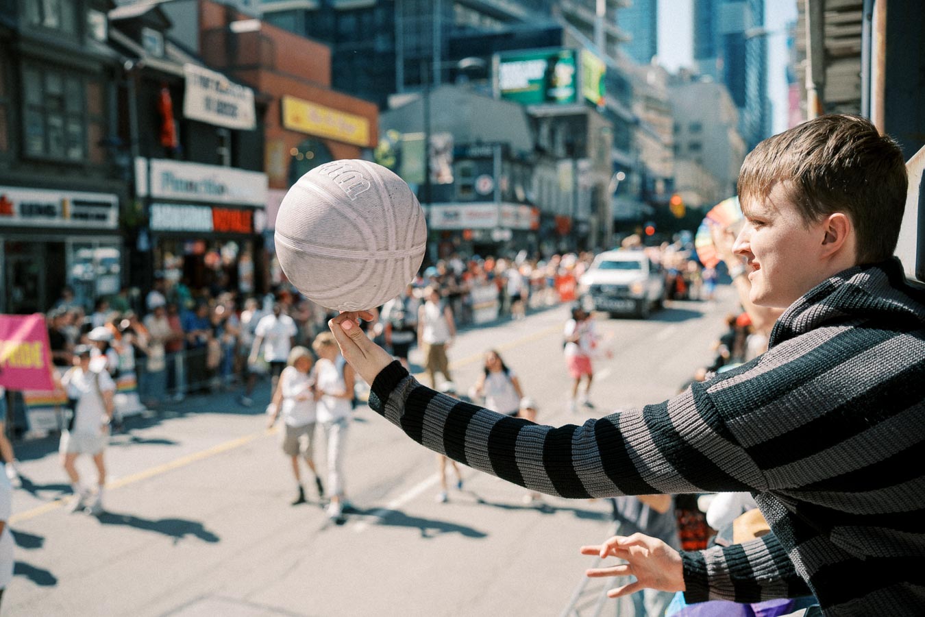 A person in a striped sweater holds a basketball while watching a lively street parade. The street is bustling with people, some carrying banners, and a white truck is visible in the background.