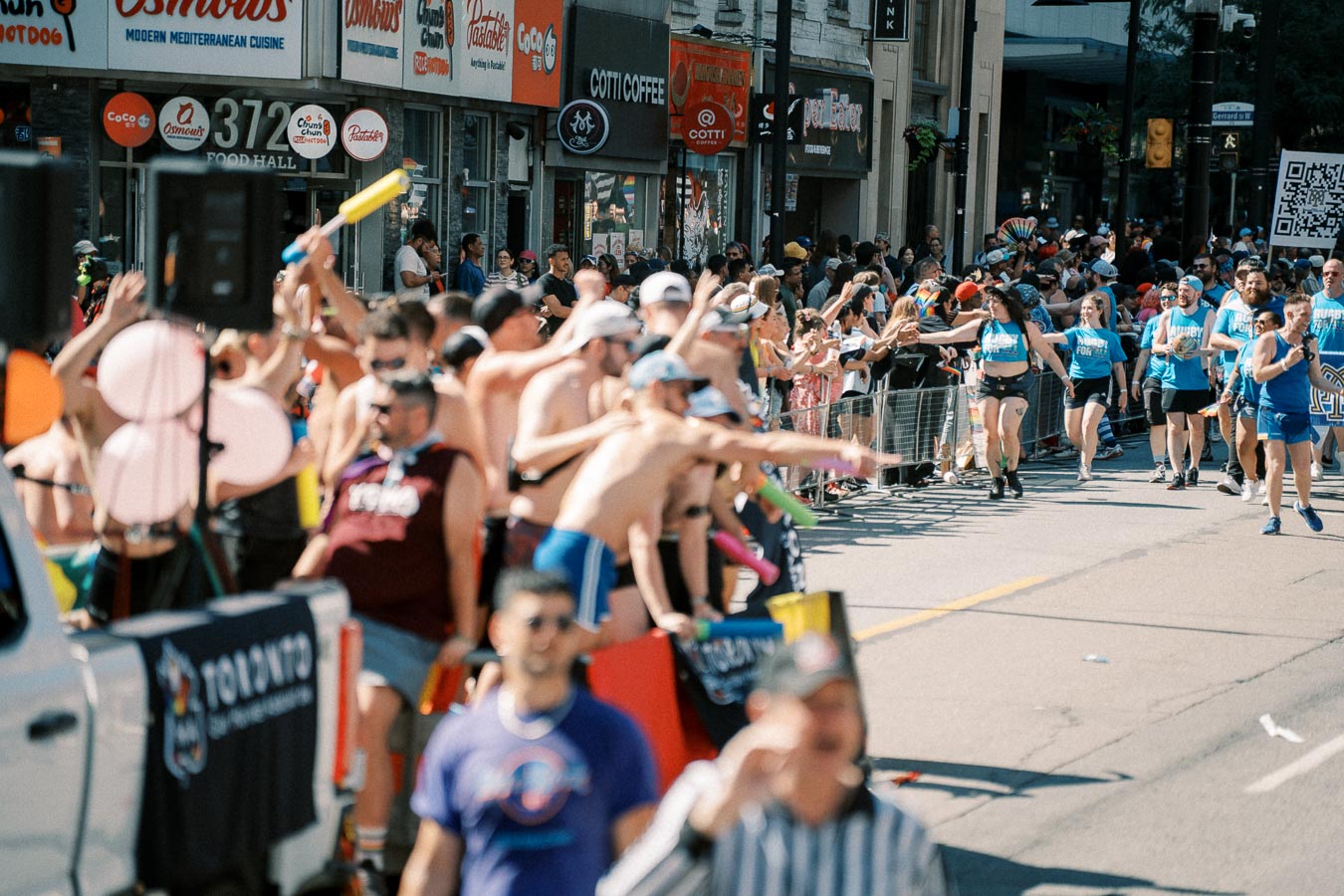 A vibrant street parade in Toronto with participants and spectators enjoying the event. People are dressed in colorful outfits, and a banner with 'Toronto' is visible, indicating the location.