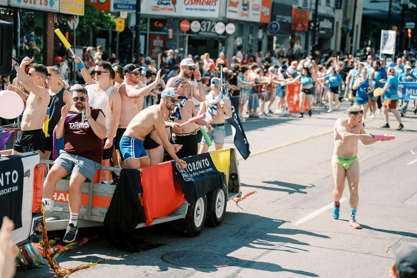 Participants in a vibrant parade, with individuals on a float and one person in bright swimwear holding a water gun, engaging with the crowd.