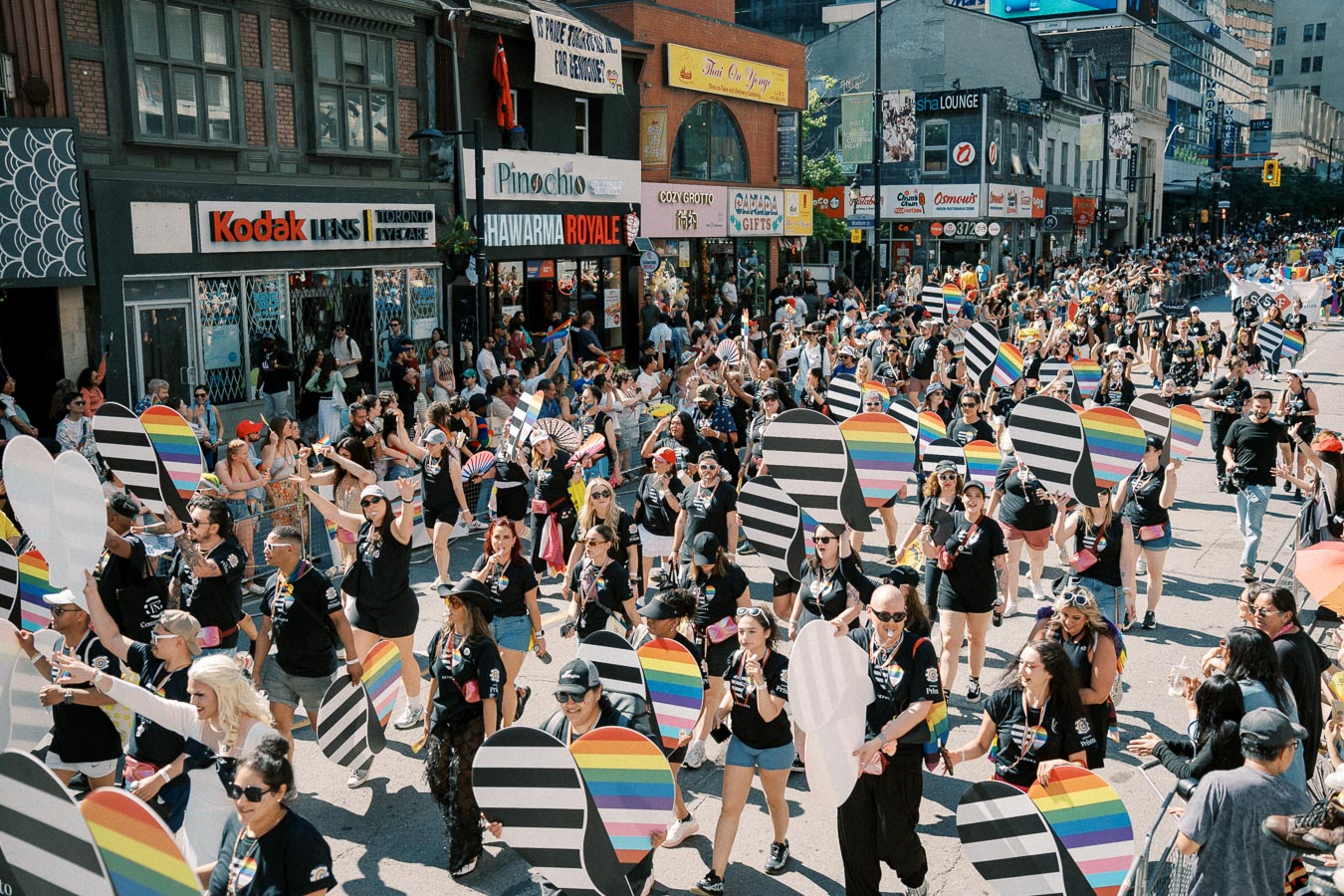 Participants in a pride parade march down a city street, holding colorful heart-shaped signs. The street is lined with spectators and various shops, creating a lively and festive atmosphere.