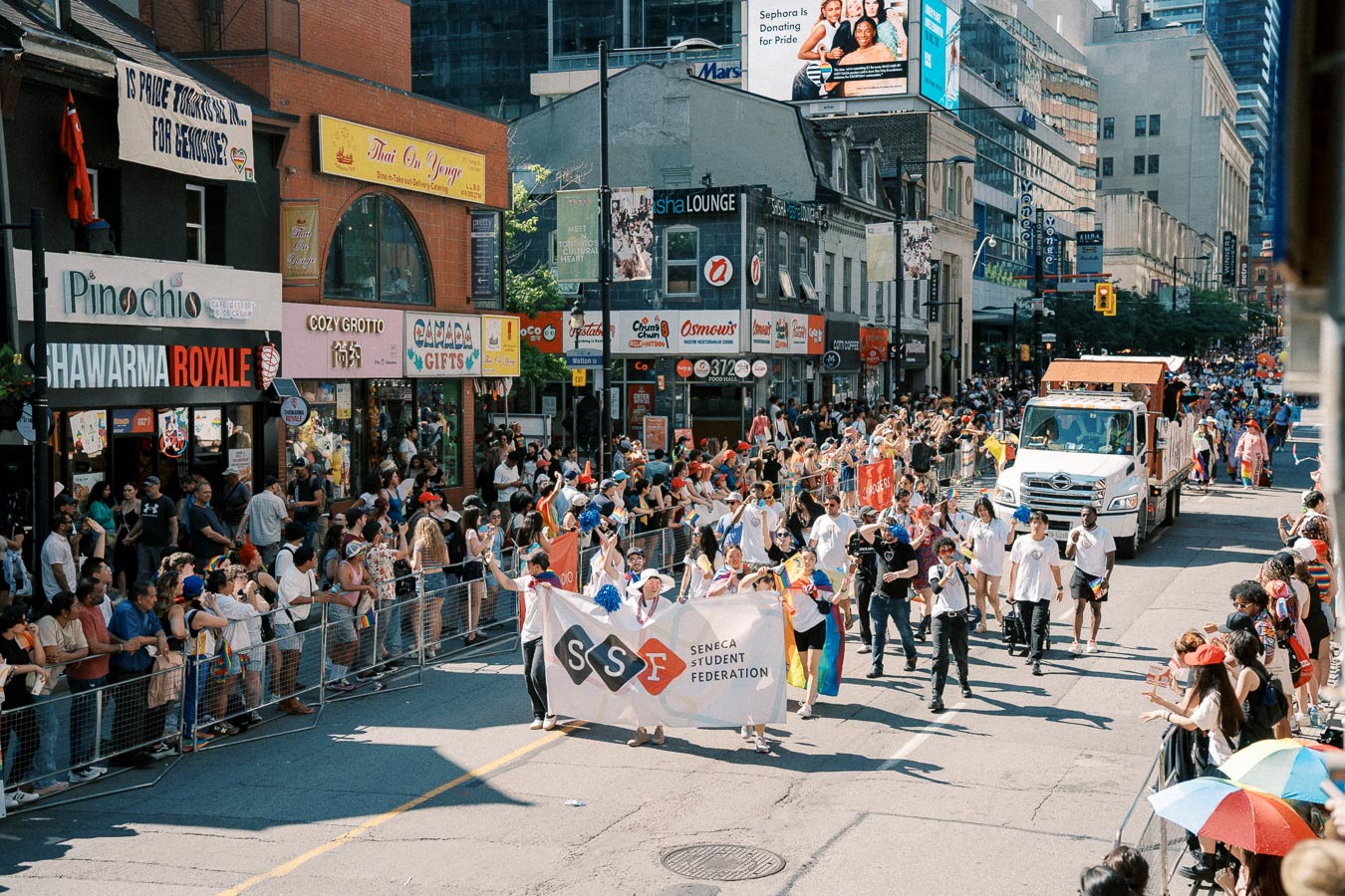 A lively parade featuring the Seneca Student Federation banner, with spectators lining the street and various shops in the background.