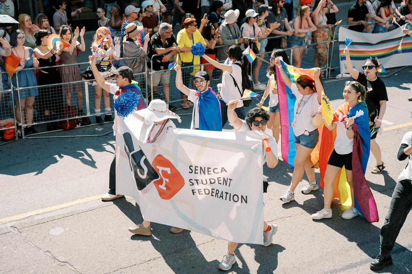 Participants from the Seneca Student Federation march in a pride parade, celebrating with rainbow flags and vibrant outfits.