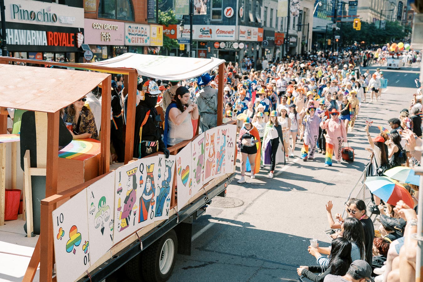 A lively parade features a float adorned with playing cards and rainbow motifs, as participants in colorful attire march down a bustling city street.