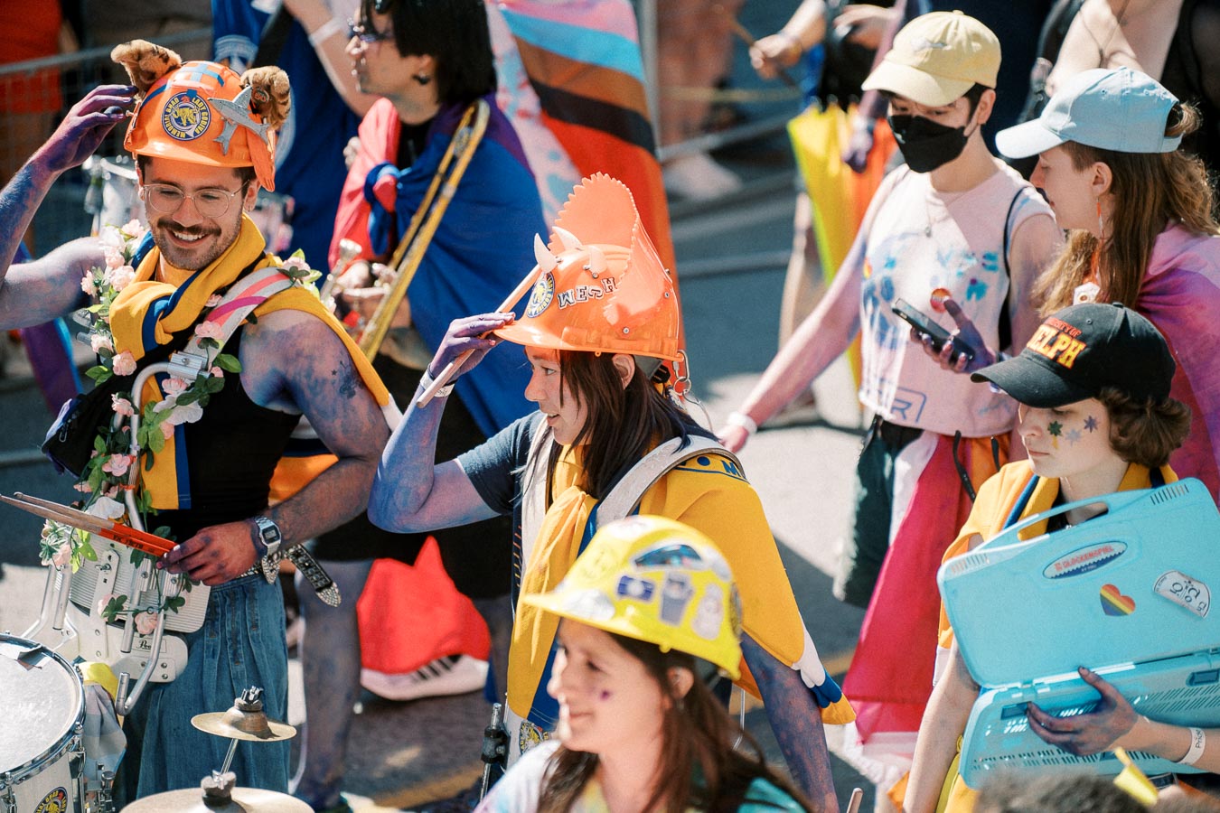 Participants in a vibrant parade wearing colorful outfits and decorated hard hats, some with painted skin and carrying musical instruments.