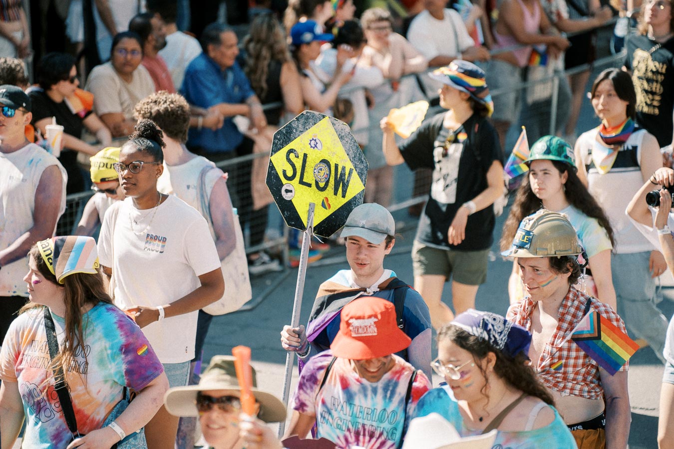 Participants in a vibrant parade, adorned with rainbow-themed attire and accessories, celebrate diversity and inclusion. A person holds a 'SLOW' sign, adding a playful touch to the festive atmosphere.