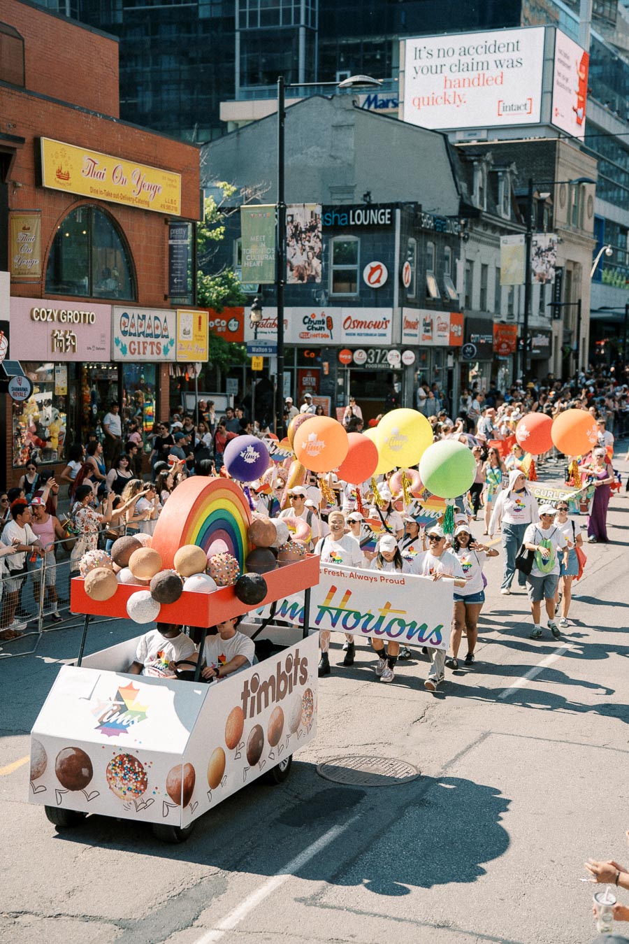 A vibrant parade featuring a Tim Hortons float with a rainbow and large Timbits, surrounded by participants holding colorful balloons.