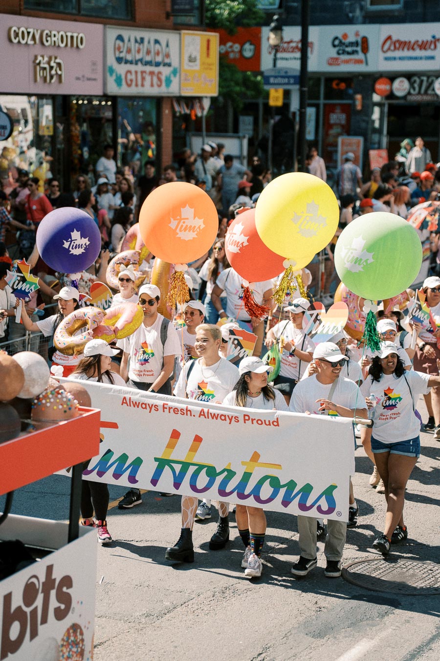 Participants in a parade hold a Tim Hortons banner and colorful balloons, celebrating with a festive crowd in the background.