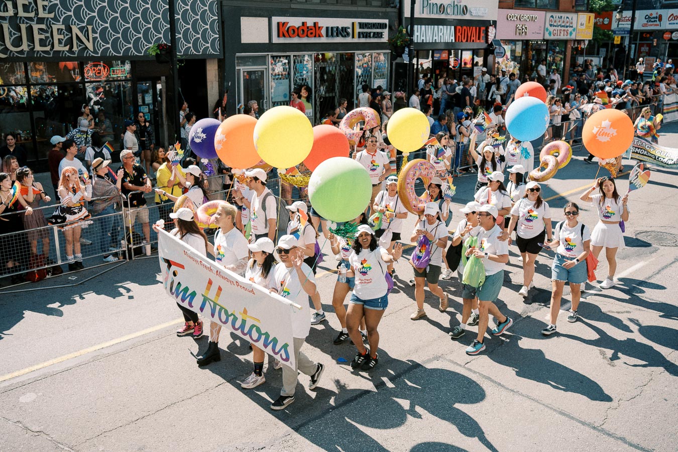 Participants in a parade hold a Tim Hortons banner and colorful balloons, celebrating with a crowd of spectators.